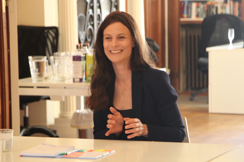 A woman sitting at a desk. She's smiling, explaining something and gesturing with her hands