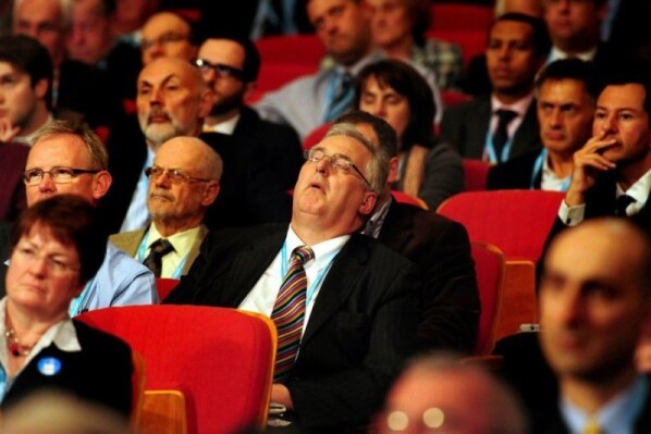 A man sleeps at a conference surrounded by attendees